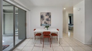 A dining area with a white table and four chairs, a painting of palm trees, and a sliding glass door leading to an outdoor area. - Costerra Naples 2BED (Naples)