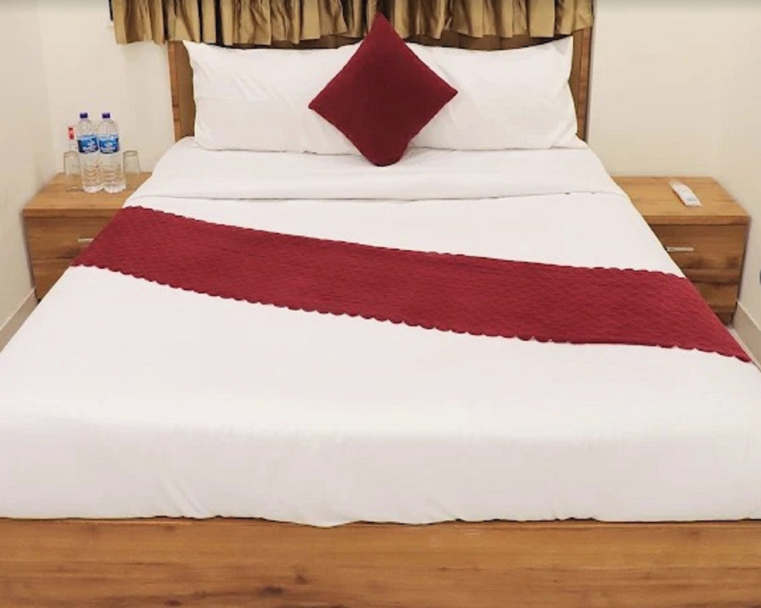 A neatly made bed with a white and red bedspread, a single red pillow, and two water bottles on a wooden nightstand.