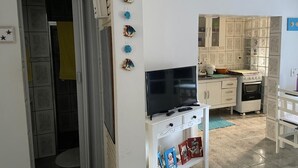 Un espacio habitable compacto con un televisor sobre un mueble blanco, una zona de cocina con alacenas y una estufa, y una puerta que da a otra habitación.