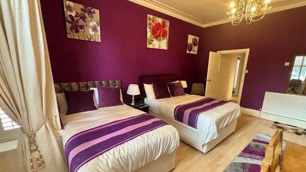 Ein Zimmer mit zwei Betten, violetter Bettwäsche, einem Kronleuchter und Kunstwerken an den Wänden.