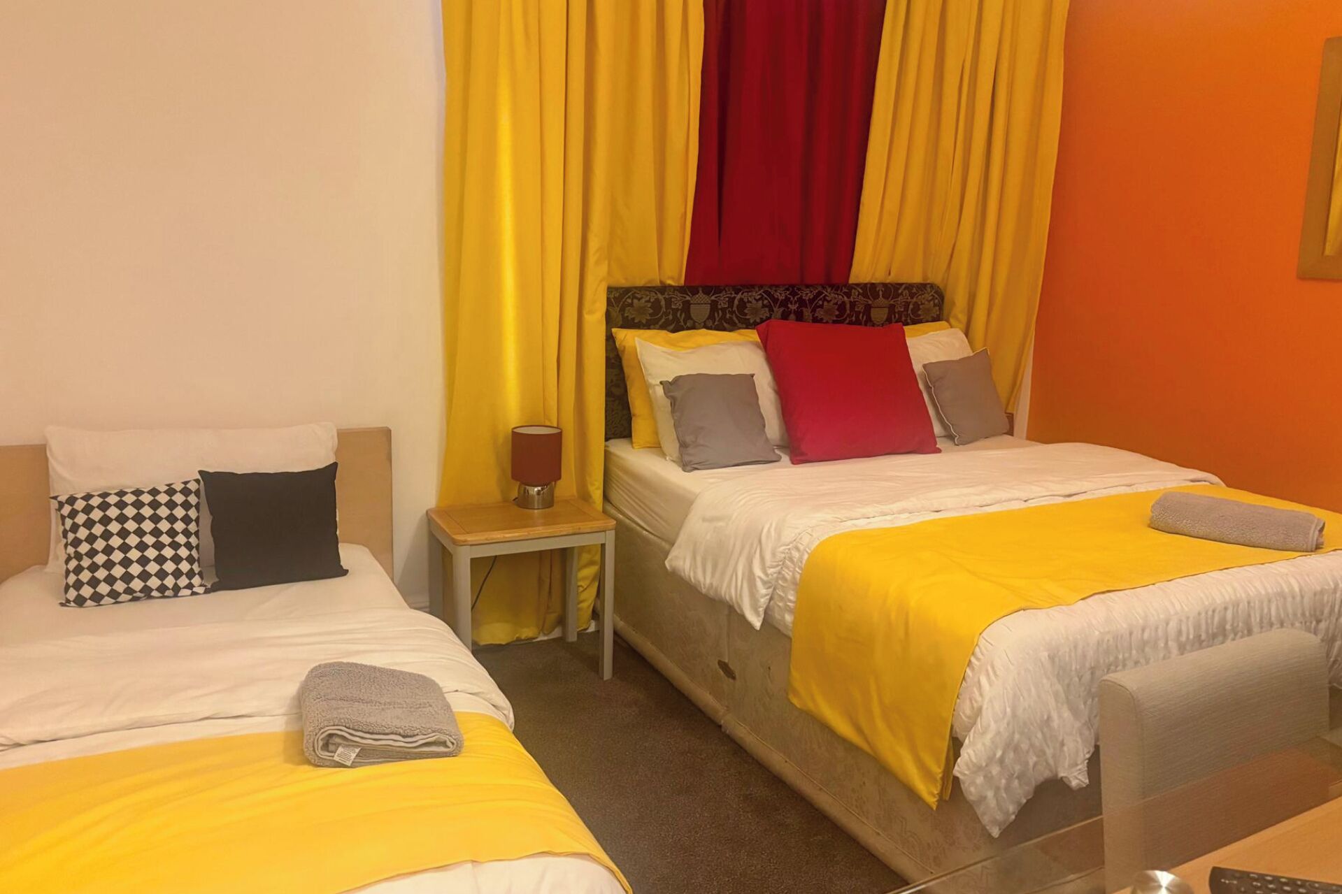 Ein Hotelzimmer mit zwei Betten, jeweils mit einer gelben Tagesdecke, einem roten Vorhang und einem kleinen Nachttisch mit Lampe.