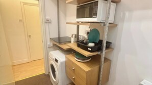 Eine kompakte Küche mit Waschmaschine, Mikrowelle und einer kleinen Arbeitsfläche mit Wasserkocher und einem grünen Teller.