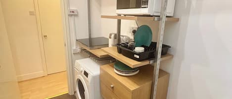 A compact kitchen with a washing machine, a microwave, and a small countertop with a kettle and a green plate.