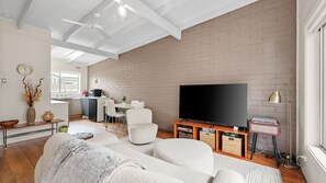 A modern living room with a flat-screen TV, a sofa, a dining area with a table and chairs, and a kitchen in the background.