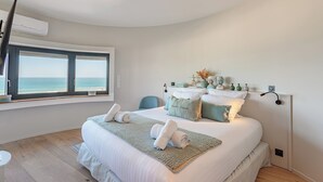 Een slaapkamer met een groot bed, uitzicht op zee door een raam, een nachtkastje en een wandlamp.