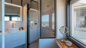 Een moderne badkamer met een grote spiegel, een ronde spiegel op een houten plank en uitzicht op de oceaan door een glazen deur.