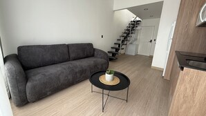 A modern living room with a grey sofa, a small round table with a plant, and a staircase leading to a white door. - Sabana Urbana (San José)