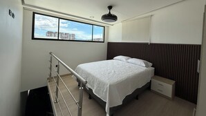 A bedroom with a bed, a nightstand, a window with a city view, a ceiling fan, and a staircase. - Sabana Urbana (San José)
