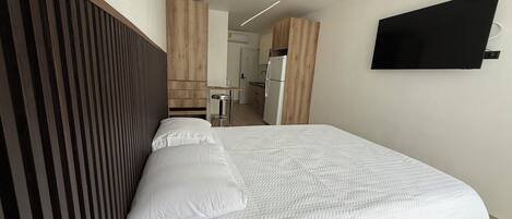 Una habitación de hotel moderna con una cama, cabecera de madera, televisor de pantalla plana y una kitchenette al fondo.
