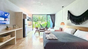 A modern hotel room with a large bed, a TV on the wall, a dining table with chairs, and a view of greenery outside.