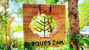 Une enseigne arborant un motif d’arbre et portant l’inscription « ARQUES ZAM », le tout dans un décor de jardin.
