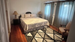 A bedroom with a bed, a chair, a nightstand, a lamp, a window with curtains, and a wooden floor. - Breathtaking villa with private pool and hot tub, 2 minute walk from the beach. (Gros Islet)