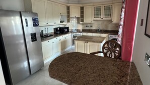 A modern kitchen with white cabinetry, a stainless steel refrigerator, granite countertops, and a dining area with a chair and table. - Breathtaking villa with private pool and hot tub, 2 minute walk from the beach. (Gros Islet)