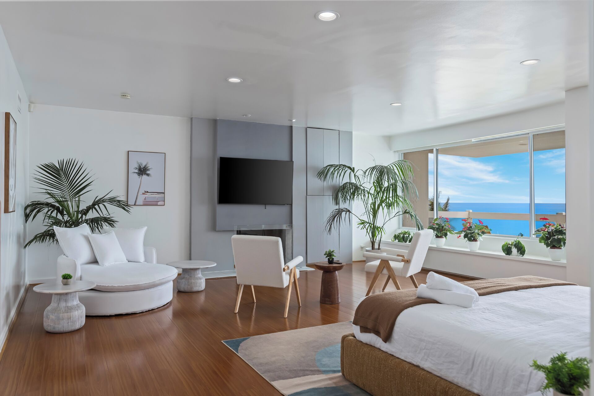 Una habitación de hotel moderna con una cama grande, un área de descanso con sillas y una mesa redonda, un televisor montado en la pared y un balcón con vistas al océano.