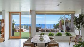 Un comedor amplio con una mesa y sillas grandes, ventanas panorámicas que ofrecen vista al océano, y un área de estar exterior bien cuidada.