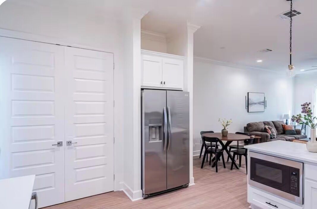 A modern kitchen with white cabinetry, a stainless steel refrigerator, a dining area with a table and chairs, and a white countertop with an oven.