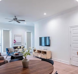 Una sala de estar moderna con una mesa de comedor redonda, un ventilador de techo, un televisor de pantalla plana montado en la pared y un mueble de madera.