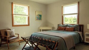 Quarto com cama, uma cadeira, uma mesinha e duas janelas com persianas.