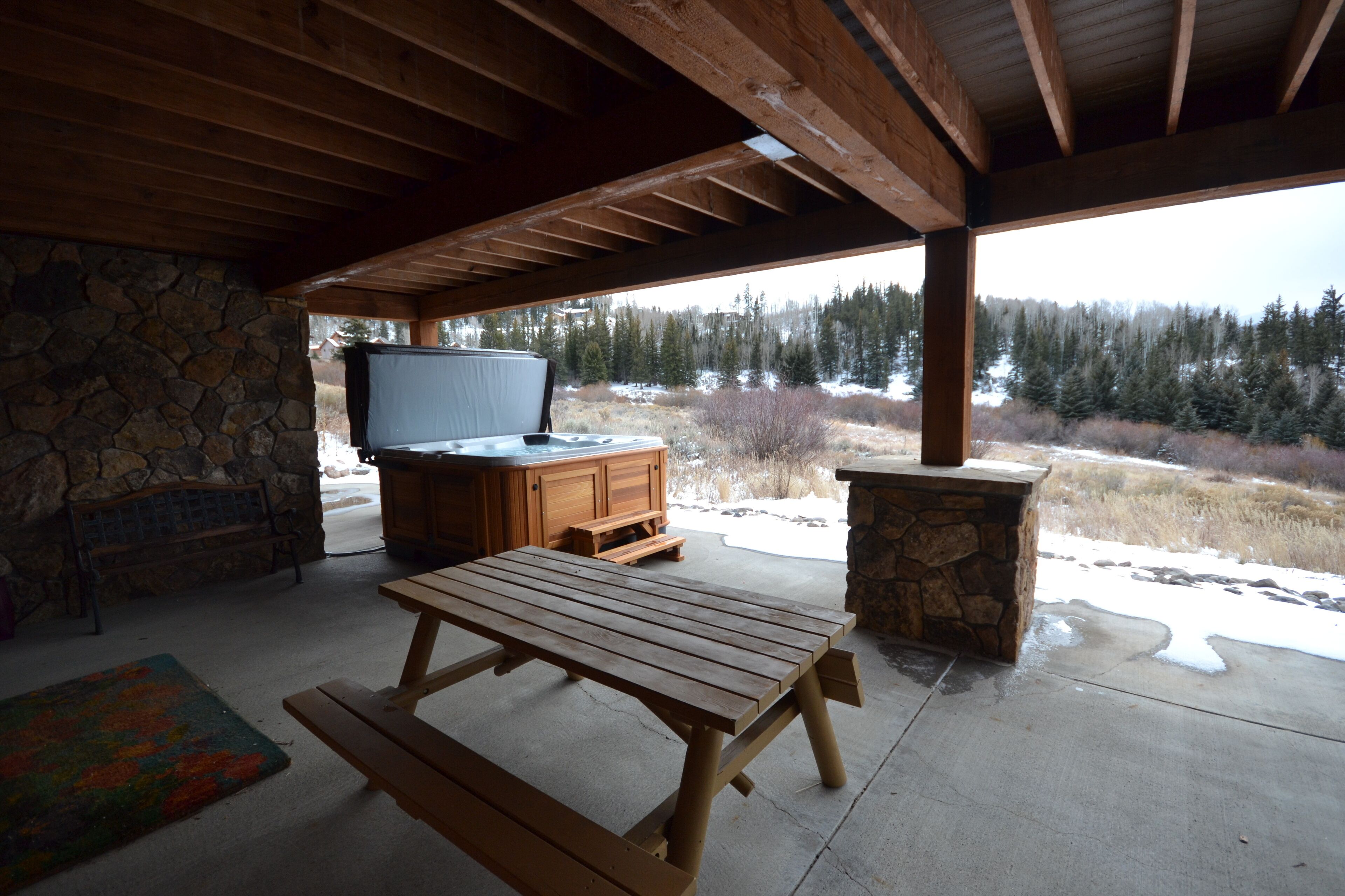 A covered outdoor area with a stone wall, a wooden table, a stone bench, and a stone fireplace.