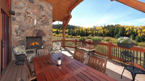 A wooden deck with a stone fireplace, outdoor seating, and a scenic view of autumn foliage. - Luxury Mountain Home with Private Hot Tub and Stunning Mountain Views (Silverthorne)