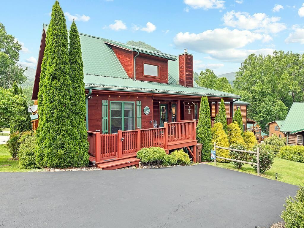 A red wooden cabin with a green roof, a porch, and a large evergreen tree in front.