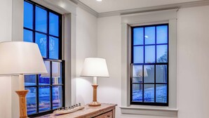 Una habitación con dos ventanas amplias, un aparador de madera con una lámpara y una mesita con adornos.