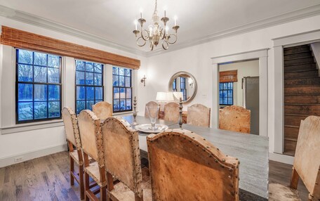 A dining room with a large table, wooden chairs, a chandelier, and large windows with blinds.