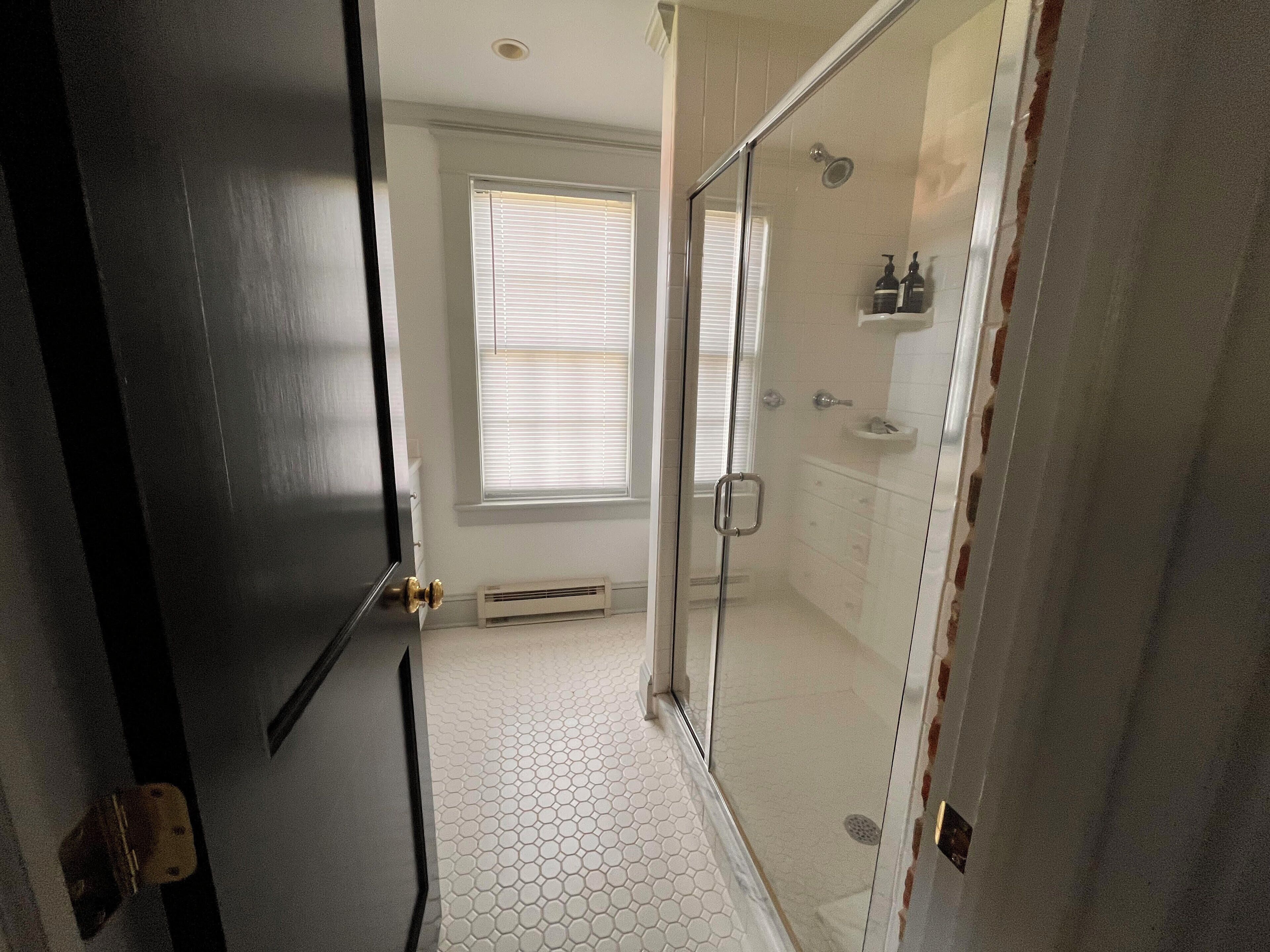 A bathroom with a glass shower enclosure, a white tiled floor, and a window with blinds.
