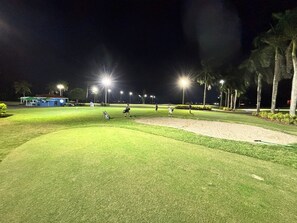 Un campo de golf de noche con luces iluminadas, palmeras y un edificio al fondo.