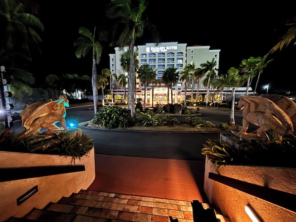 Entrada de hotel con palmeras, una estatua de una criatura y un cartel que dice 'EMBASSY SUITES'.