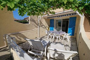 A patio with white plastic chairs and a table, a blue shuttered door, and a tree providing shade.