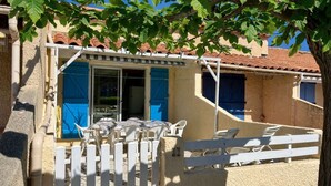 A covered outdoor seating area with a table and chairs, a white fence, and a building with blue shutters. - Air-conditioned holiday home for 5 people, secure residence MAS DE LA PLAGE, south-facing... (PORTIRAGNES)