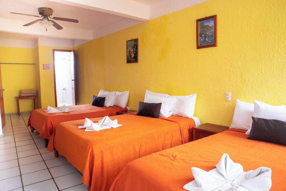 A hotel room with three beds, each with orange bedding and white pillows. There is a ceiling fan, a small table, and a painting on the wall.