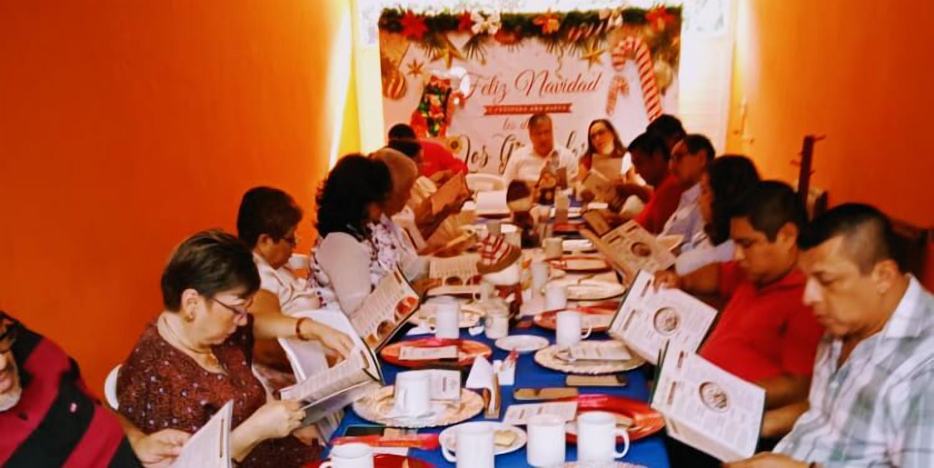 Un grupo de personas sentadas alrededor de una mesa con platos, tazas y libros, en una habitación con un ambiente festivo.
