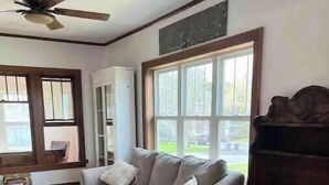 A living room with a sofa, a piano, and a window with a view of a yard. - Restyle Farm house (Baldwin)