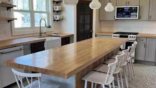 A modern kitchen with a long island, white bar stools, pendant lighting, and a TV mounted above the counter. - Restyle Farm house (Baldwin)