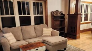 A living room with a beige sectional sofa, a small white side table, and a wooden coffee table. There is a dark wooden bookshelf and a wooden cabinet in the background. - Restyle Farm house (Baldwin)