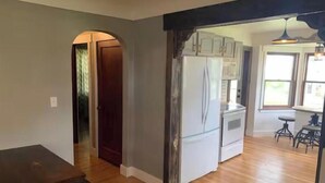 Eine Küche mit weißen Schränken und einem Kühlschrank, ein Essbereich mit einem Holztisch und ein Blick ins Freie durch ein Fenster.