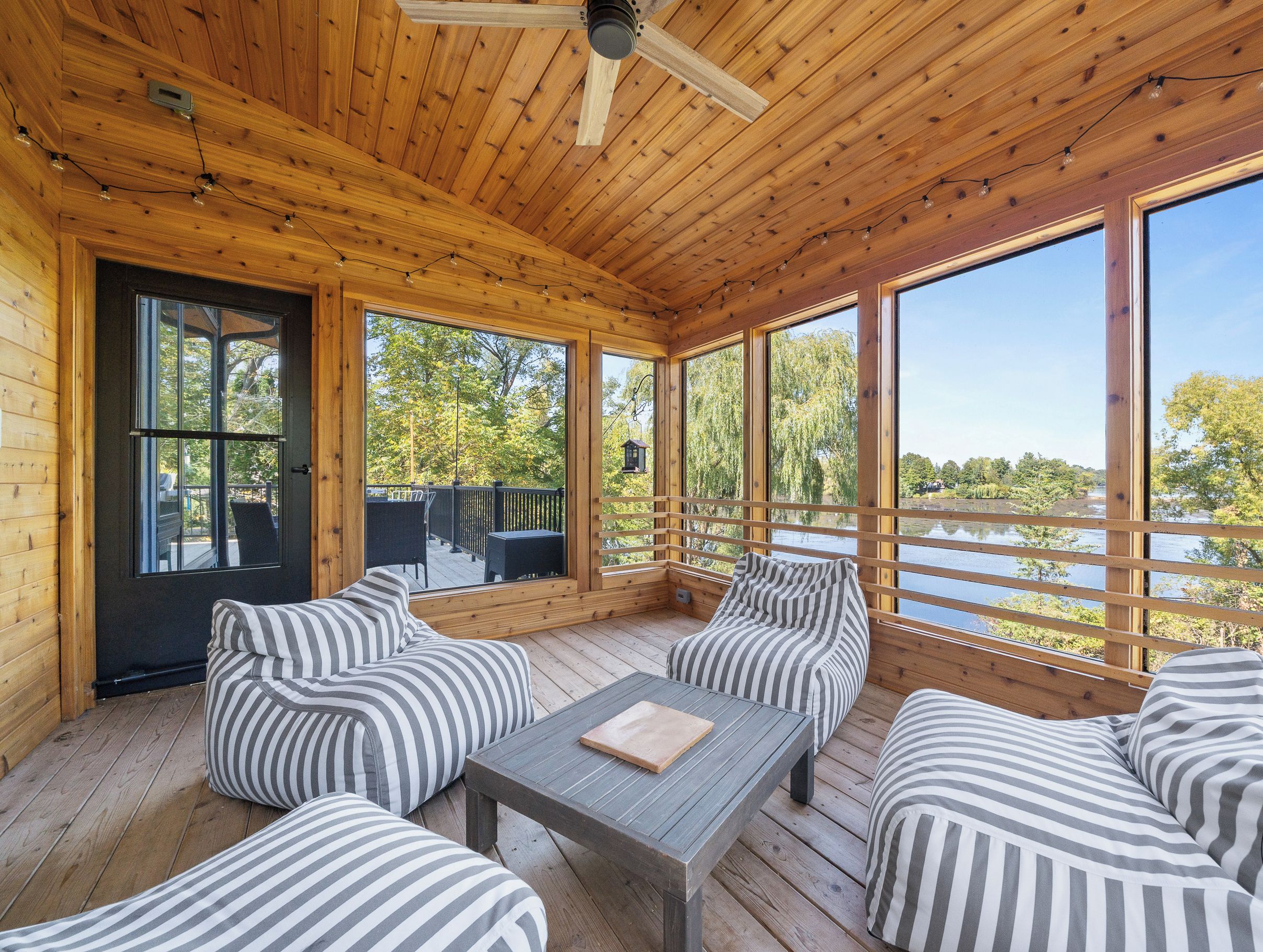 A wooden deck with striped cushions and a table, large windows offering a view of trees and a body of water.