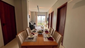 A dining area with a wooden table set for four, chairs, and a view of a balcony with a plant. - Apto Pet Friendly com Piscina a 100m da Orla (Japaratinga)