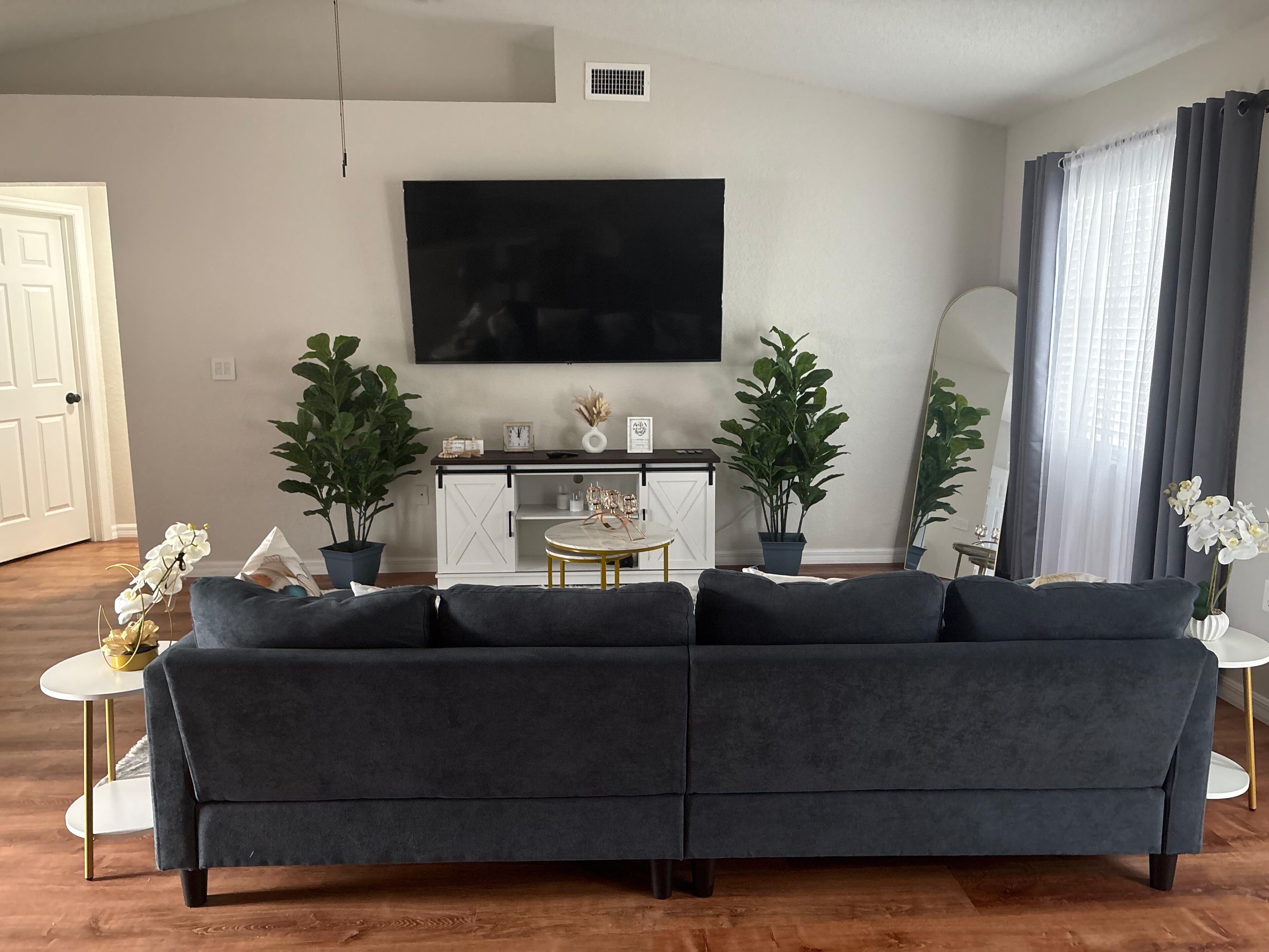 Un salon avec un canapé d’angle gris foncé, un meuble blanc, un téléviseur à écran plat fixé au mur et deux plantes en pot.