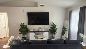 Un salon avec un canapé d’angle gris foncé, un meuble blanc, un téléviseur à écran plat fixé au mur et deux plantes en pot.