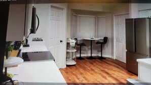 Une cuisine compacte avec des meubles blancs, un plancher en bois, un coin repas comprenant une petite table et des tabourets de bar, et un réfrigérateur.