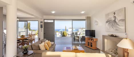 A modern living room with a sofa, coffee table, and TV, a dining area with chairs and a table, and a balcony with a view of the sea.