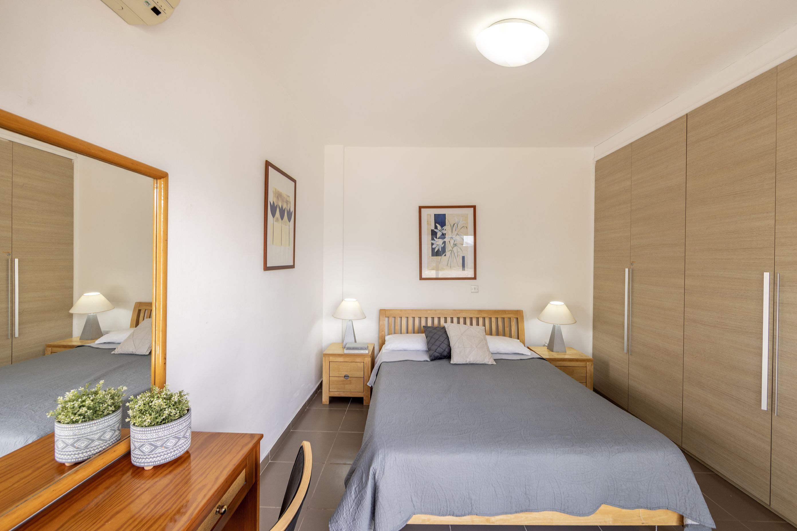 A bedroom with two beds, wooden wardrobe, bedside tables with lamps, and a mirror.