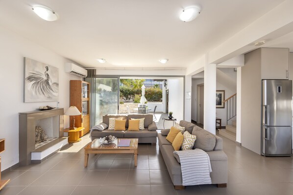 A spacious living area with a sofa set, a coffee table, and a fireplace. There is a large window providing a view of the outdoors.