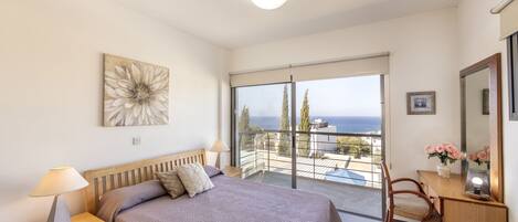 A bedroom with a bed, bedside tables, a desk, and a chair. There is a balcony with a view of the sea.