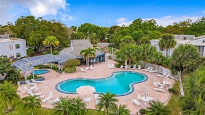 A large outdoor swimming pool surrounded by lounge chairs and palm trees, with a covered seating area and a view of the surrounding greenery. - Upstay 2BR Family Condo Ventura Golf Country Club (Orlando)