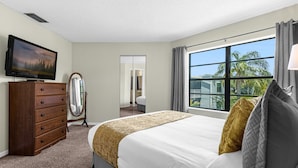 A hotel room with a large bed, a wooden dresser, a flat-screen TV, a mirror, and a window with a view of palm trees. - Upstay 2BR Family Condo Ventura Golf Country Club (Orlando)