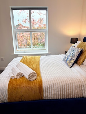 Uma cama bem arrumada com manta branca e dourada, duas toalhas brancas dobradas e vista para árvores de outono pela janela.